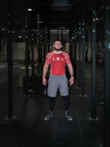 Szeroki kadr przedstawiający mężczyznę stojącego dumnie w metalowej bramie do przysiadów (squat rack) w nowoczesnej, ciemnej siłowni. Fotografia reklamowa odzieży sportowej Polska.