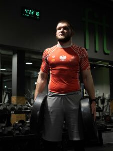 Bliższe ujęcie muskularnego mężczyzny chwytającego czarne hantle na stojaku na siłowni, w czerwonej polskiej koszulce zapaśniczej. Sesja sportowa w Łodzi.