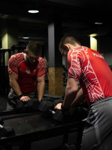 Muskularny mężczyzna w czerwonej koszulce z polskim orłem i godłem na siłowni, stojący przy stojaku z czarnymi hantlami i patrzący w dół.