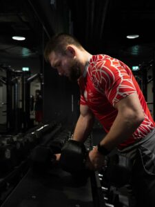 Muskularny mężczyzna w czerwonej koszulce z polskim orłem i godłem na siłowni, stojący przy stojaku z czarnymi hantlami i patrzący w dół.