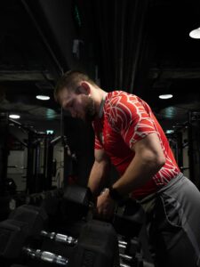 Muskularny mężczyzna w czerwonej koszulce z polskim orłem i godłem na siłowni, stojący przy stojaku z czarnymi hantlami i patrzący w dół.