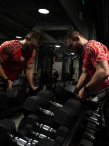 Muskularny mężczyzna w czerwonej koszulce z polskim orłem i godłem na siłowni, stojący przy stojaku z czarnymi hantlami i patrzący w dół.