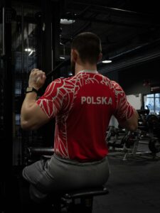 Widok z tyłu na mężczyznę trenującego plecy na maszynie do ściągania drążka na siłowni. Widoczny napis POLSKA na czerwonej koszulce sportowej. Trening siłowy.