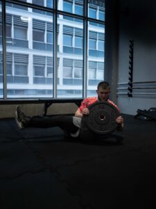 Mężczyzna wykonuje ćwiczenie "Russian twist" (skręty tułowia) z ciężkim talerzem obciążeniowym na podłodze siłowni. Tło z dużym oknem. Sesja wizerunkowa sportowca.