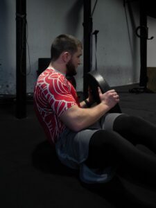 Mężczyzna wykonuje ćwiczenie "Russian twist" (skręty tułowia) z ciężkim talerzem obciążeniowym na podłodze siłowni. Tło z dużym oknem. Sesja wizerunkowa sportowca.