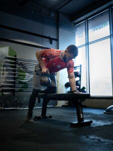 Mężczyzna w polskiej koszulce zapaśniczej wykonuje wiosłowanie kettlebell jednorącz w podparciu o ławkę treningową na siłowni, chwytając ciężki kettlebell.