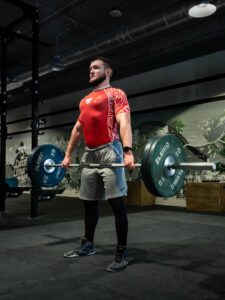 Mężczyzna w polskiej koszulce sportowej z orłem i godłem stoi wyprostowany, trzymając sztangę Eleiko z zielonymi talerzami po wykonaniu martwego ciągu na siłowni.