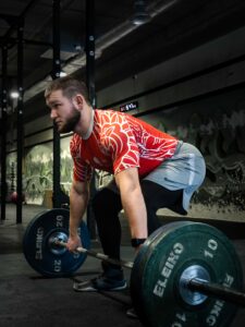 Mężczyzna w polskiej koszulce sportowej z orłem i godłem stoi wyprostowany, trzymając sztangę Eleiko z zielonymi talerzami po wykonaniu martwego ciągu na siłowni.