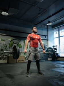 Sportowiec w czerwono-białej koszulce sportowej z napisem POLSKA trzyma sztangę Eleiko z zielonymi talerzami. Fotografia wykonana w Crossfit boxie w Łodzi.