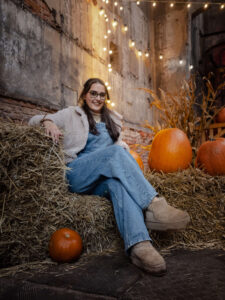 Uśmiechnięta kobieta w dżinsowych ogrodniczkach siedząca na snopkach siana wśród dyń. Jesienna sesja portretowa w industrialnym wnętrzu Fuzji z ciepłym oświetleniem. Fotograf Łódź.