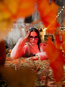 Zbliżenie na twarz kobiety w jasnej kurtce, opierającej się o siano. Jesienny portret sfotografowany przez rozmyte, pomarańczowe liście na pierwszym planie. Sesje kobiece Łódź.