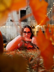 Portret kobiety w okularach opierającej głowę na dłoniach, sfotografowany przez rozmyte jesienne liście. Sesja zdjęciowa na tle snopków siana i ciepłych żarówek w industrialnym wnętrzu. Fotograf portretowy Łódź.