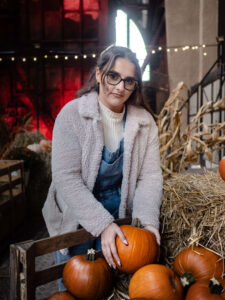 Uśmiechnięta kobieta w jasnej kurtce i ogrodniczkach układająca pomarańczowe dynie w drewnianej skrzyni. Jesienna sesja portretowa z elementami rustykalnymi. Profesjonalna fotografia kobieca Łódź.