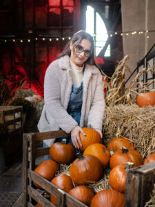 Kobieta w okularach patrząca w obiektyw, stojąca przy drewnianej skrzyni pełnej pomarańczowych dyń. Klimatyczna sesja zdjęciowa w pofabrycznym klimacie z czerwonym podświetleniem. Fotograf Łódź.