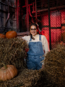 Kobieta w okularach oparta o snopki siana z pomarańczowymi dyniami. W tle pofabryczna siatka i czerwone oświetlenie. Klimatyczna kobieca sesja zdjęciowa w Łodzi.
