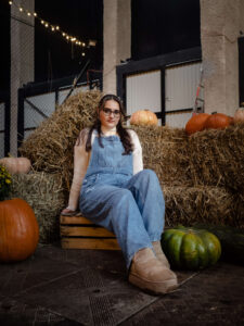 Kobieta siedząca na drewnianej skrzynce obok zielonych i pomarańczowych dyń na tle siana i ceglanego muru. Naturalny portret jesienny, sesja fotograficzna Łódź.