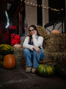Radosna kobieta siedząca wśród jesiennych dekoracji – dyń, siana i wrzosów. Rustykalna sesja portretowa zrealizowana w pofabrycznych przestrzeniach Łodzi.
