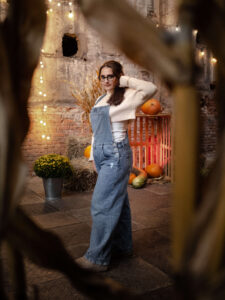 Kobieta pozująca w ogrodniczkach na tle jesiennej scenografii i wiszących światełek. Zdjęcie portretowe z kreatywnym rozmyciem pierwszego planu. Profesjonalny fotograf Łódź.